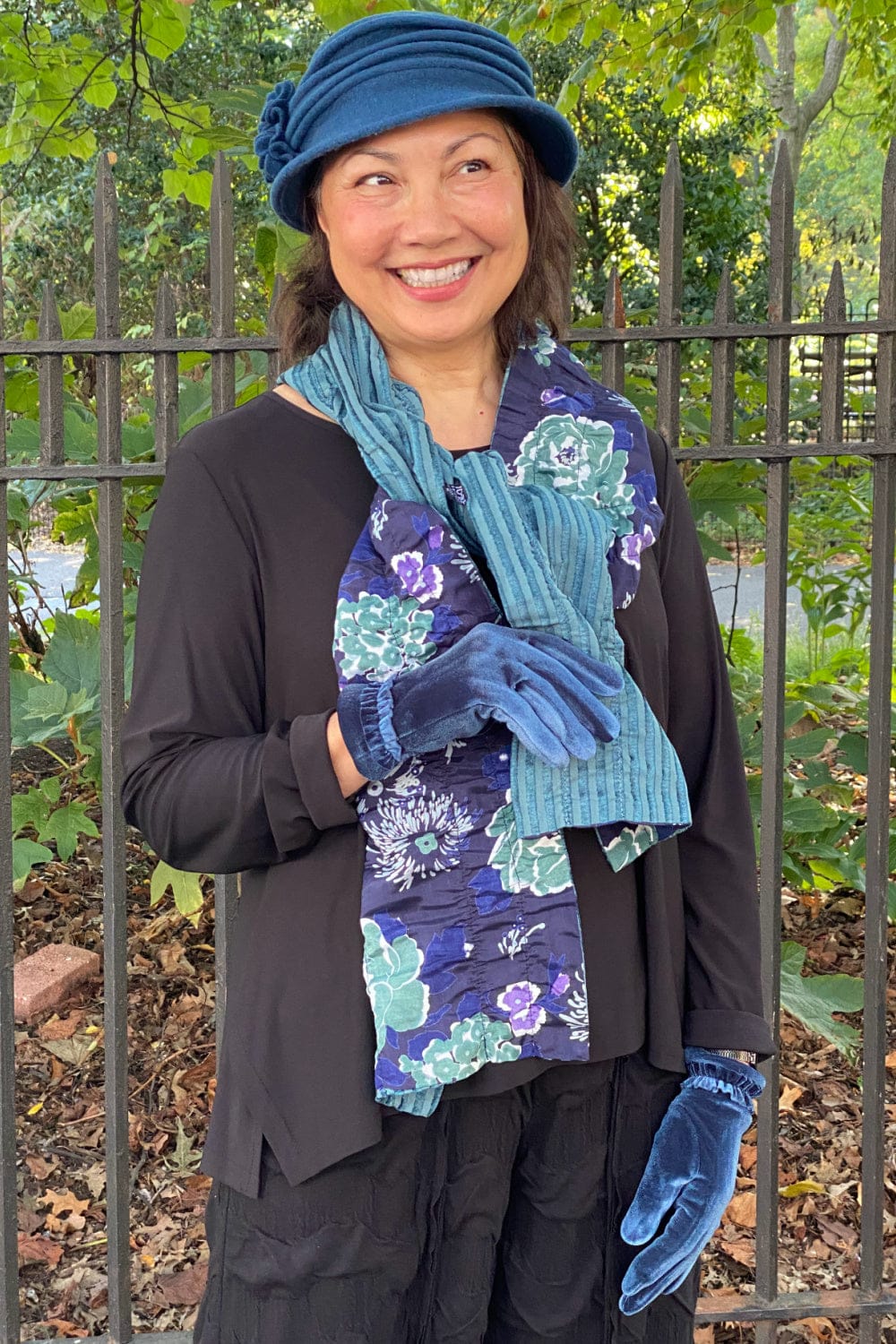 Happy woman standing in a park wearing women's winter accessories. Blue small rim hat, reversible velvet blue floral scarf and blue velvet gloves.