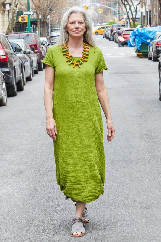 Older woman with long grey hair wearing a moss green short sleeve long cotton dress. She is also wearing beige sandles