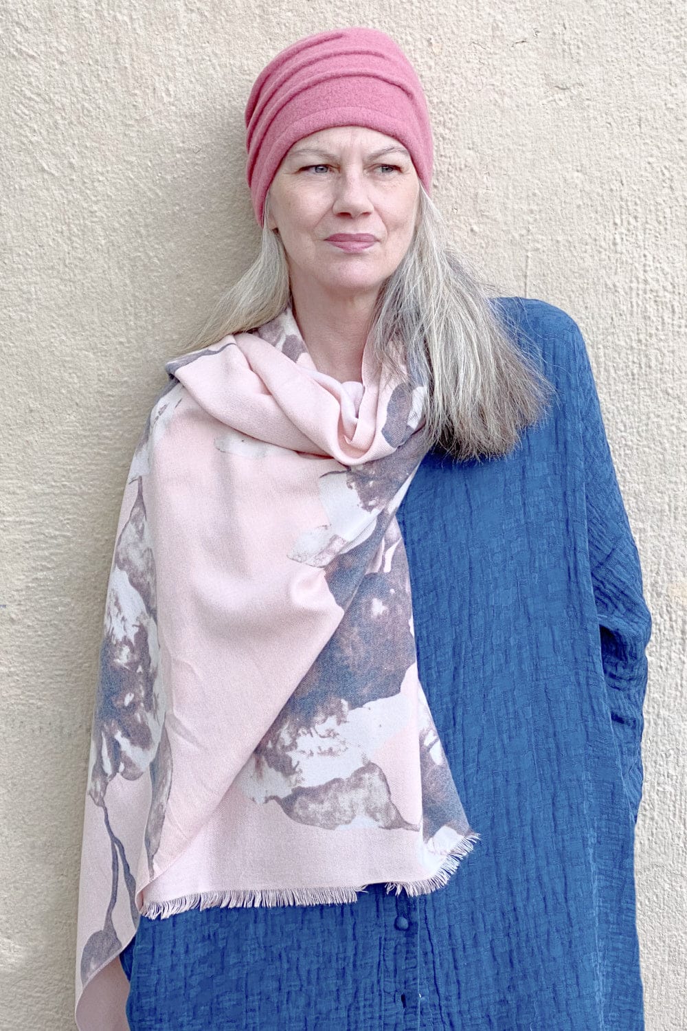 Soft pink scarf with grey floral print being worn by a woman with long grey hair. She  is wearing a pink snug wool hat.