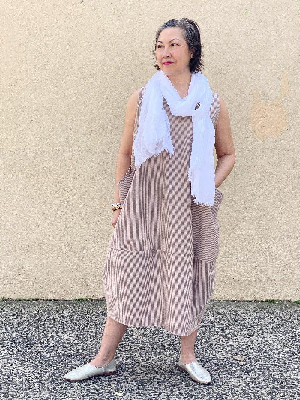 Woman wearing a seersucker white and mocha stripe tank dress with two front pockets and white summer scarf.