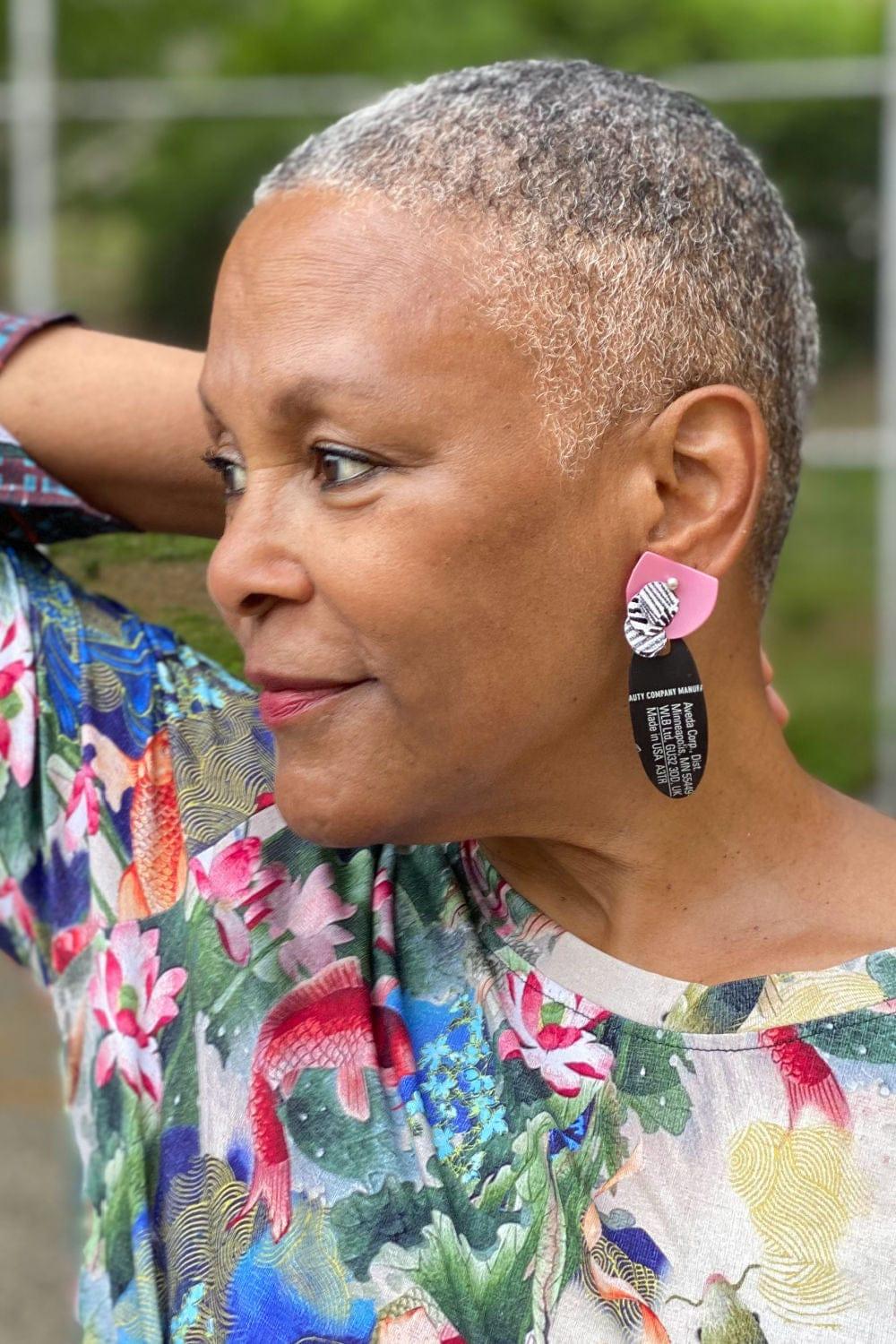 Cropped haired woman wearing fun recycled plastic earrings made up of shampoo bottles. They are black, pink and white.