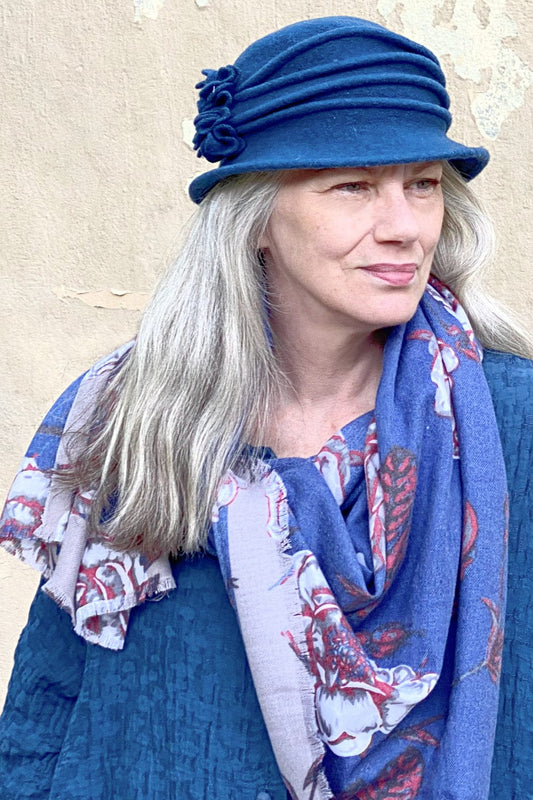 Womens Merino Wool hat with pleat detail and 2 side flowers. Grey haired model is wearing a floral blue scarf.