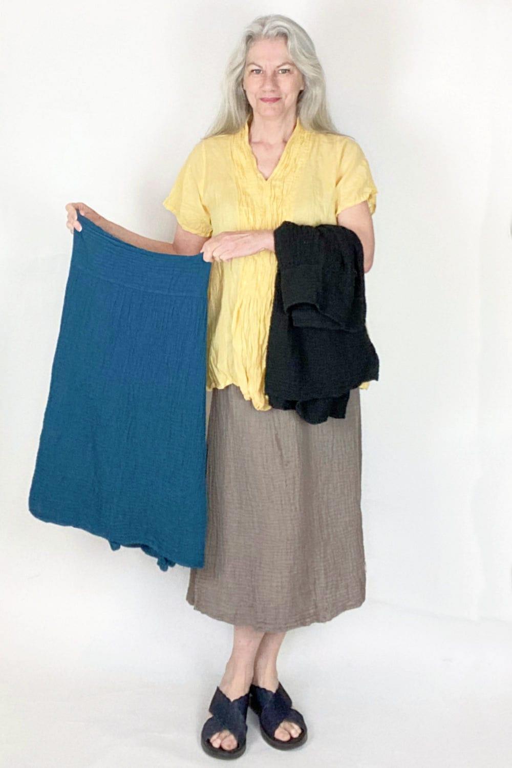 Woman with long grey hair wearing a yellow cotton short sleeve cotton blouse with a beige cotton skirt. She is holding a blue  skirt in her hands and has a black skirt draped over her arm.