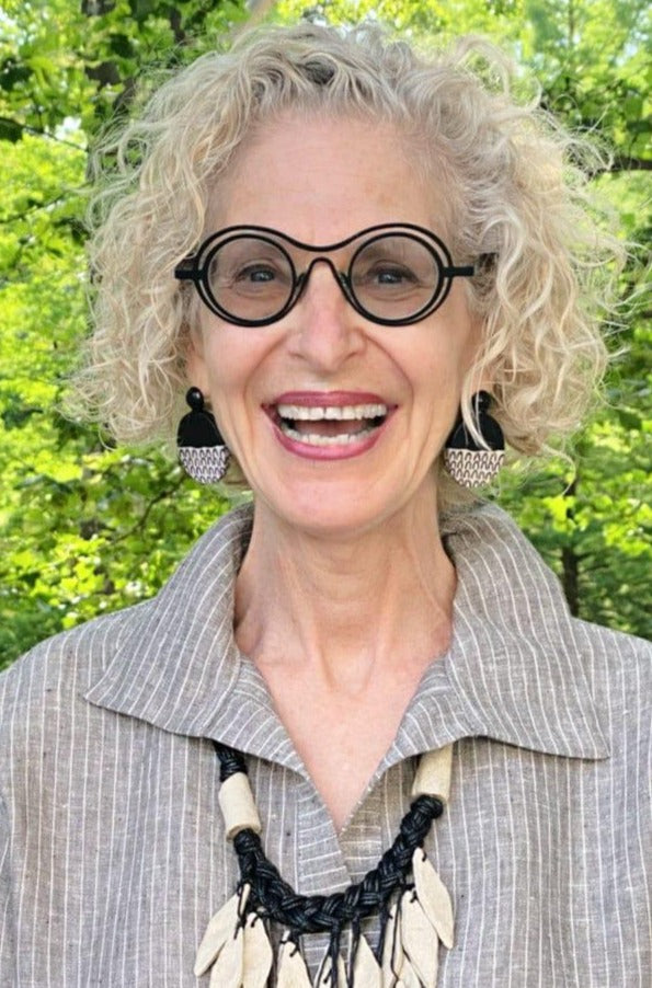 Blond older woman smiling, wearing black and white earrings with a necklace made of recycled material. She is wearing a linen grey and white pinstripe tuni dress.