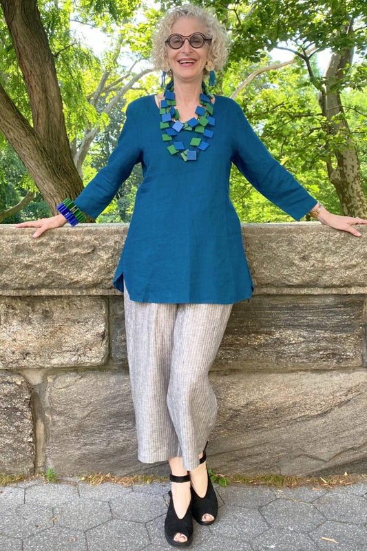 Pretty woman wearing a teal linen long sleeve top with grey linen pants. She is wearing wooden necklaces and bracelets colored green and blue.