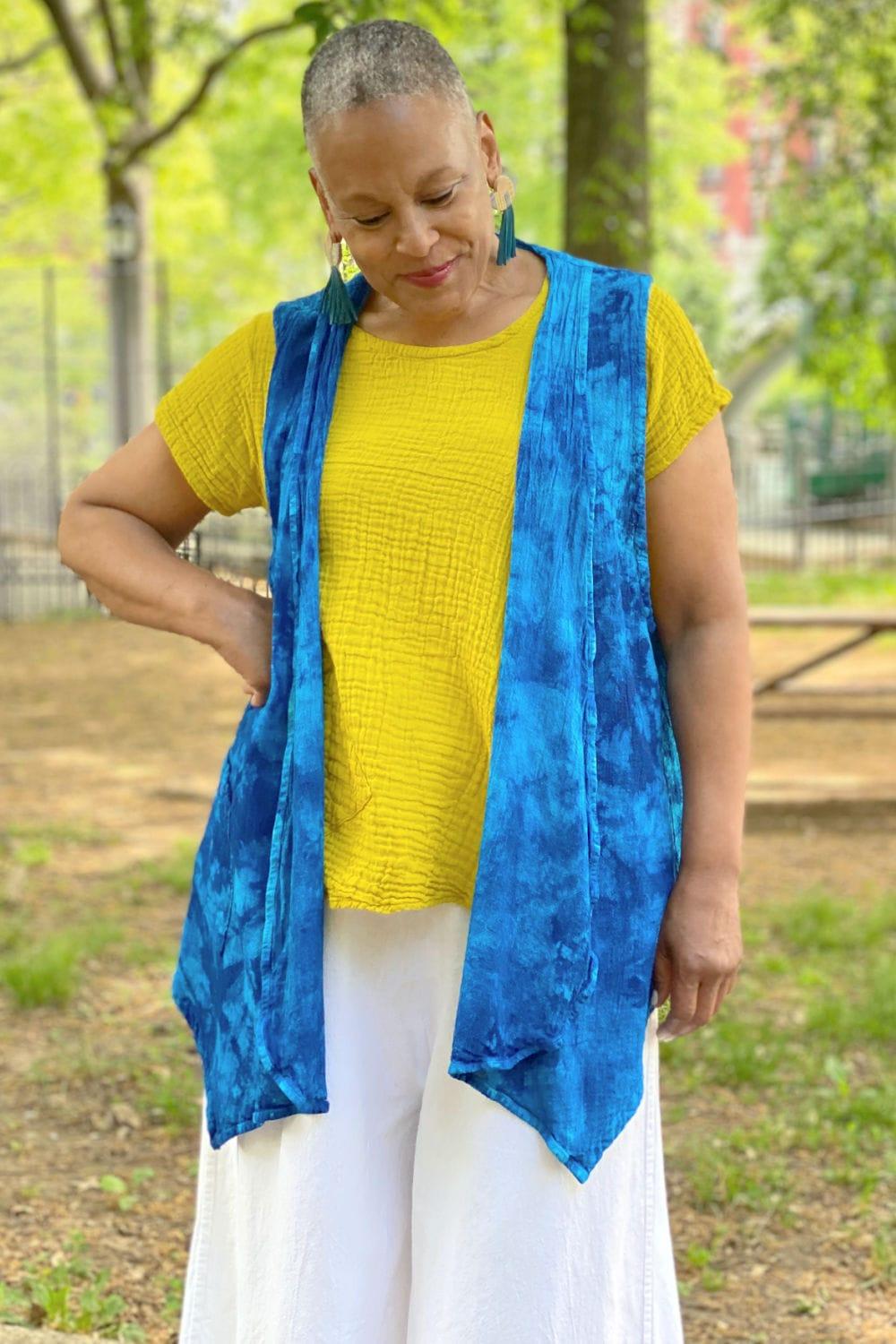 Women wearing a bright blue cotton vest overtop a yellow cotton shortsleeve tee and white wide leg pants. 