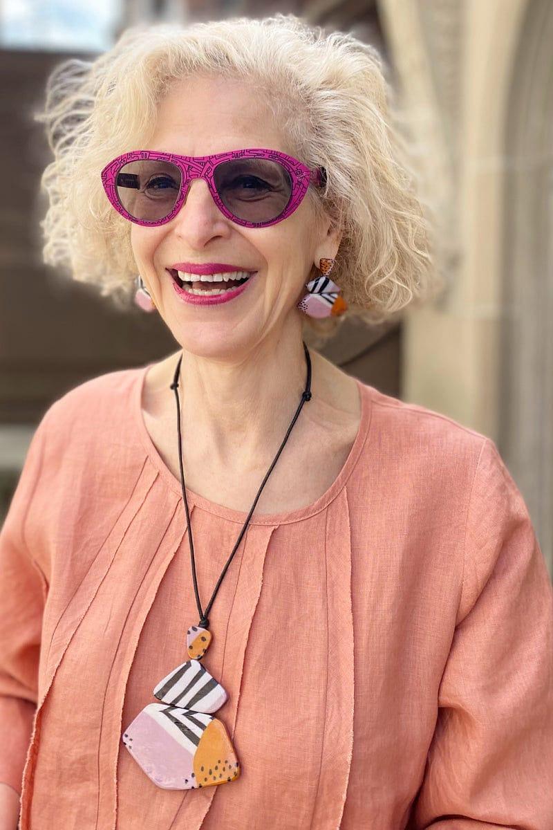 Smiling Older woman with blond hair wearing pink glasses wearing peach linen top with decorative stitching. She is wearing a Three tier wooden pendant neckacle with black and white stripes and dots. Flat wooden beads stacked from small to large and strung on an adjustable black cord. She is also wearing matching earrings.