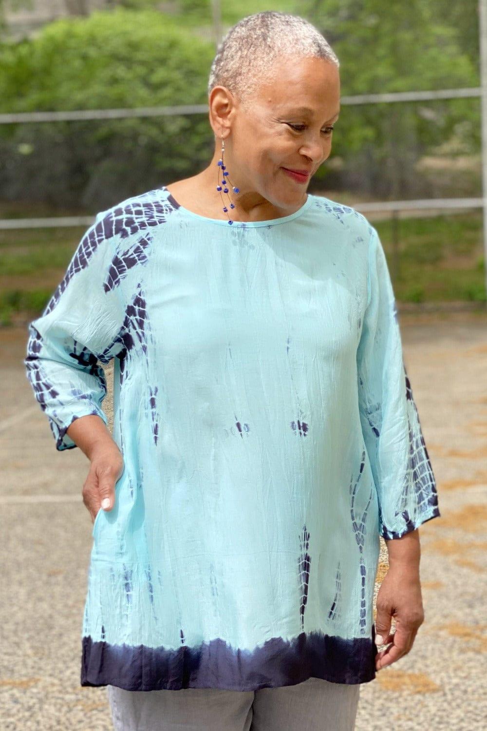 Woman wearing a tie dye Aline shaped top in blue hues. She is also wearing dangle wooden earrings and natural color linen pants.
