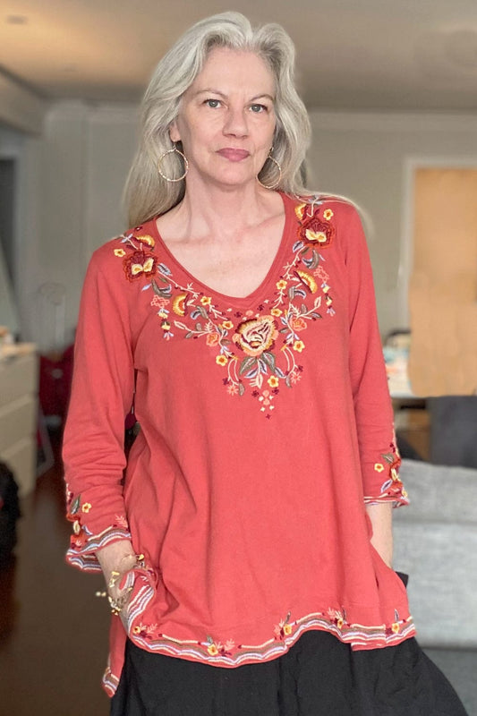 Woman with long grey hair wearing a very pretty coral colored v neck top with floral embroidery.