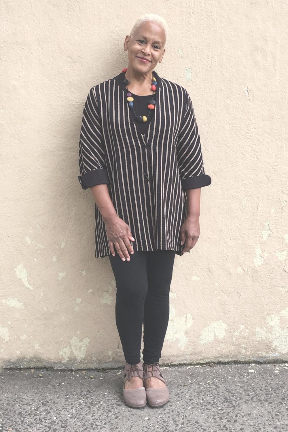 Women's Black Stripe Jacke is being worn by a smiling older woman resting against a wall. She is also wearing black leggings and a multicolored single strand necklace. She is smiling.