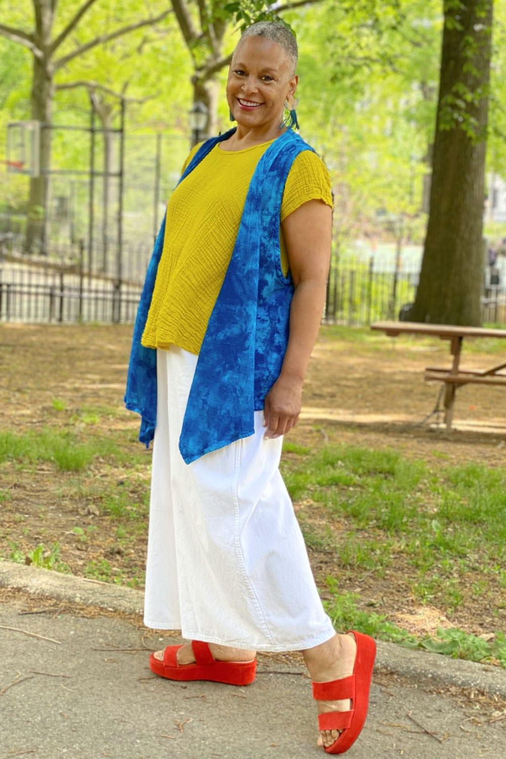 Smiling older woman wearing a bright yellow tee with a blue cotton vest and full leg white pants.