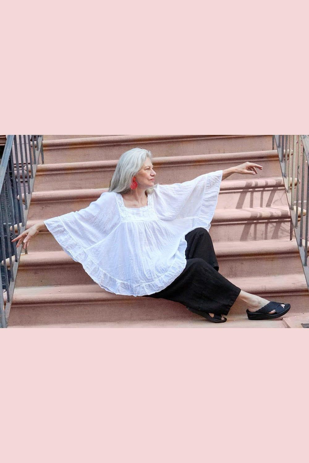 Woman wearing a curved white cotton blouse with red earrings and black linen full cut pants sitting outside on stairs.