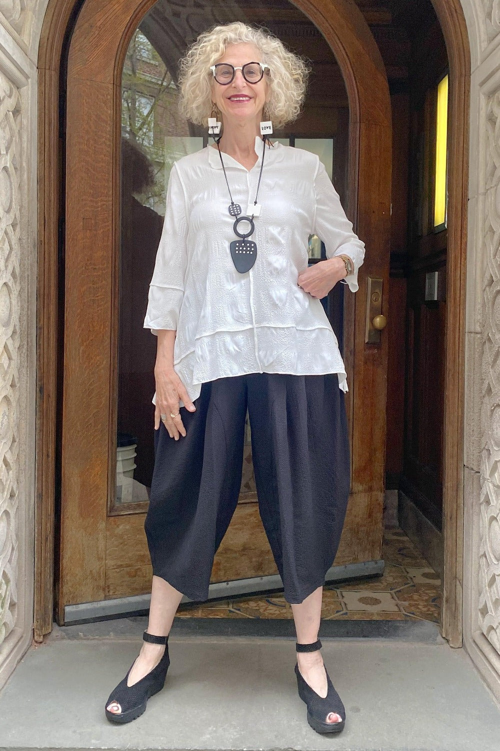 White Aline Shape top with  black crop bubble cut pant being worn on a smiling blond woman. She is standing in a doorway outsite. She is also wearing a black and white necklacke and earrings