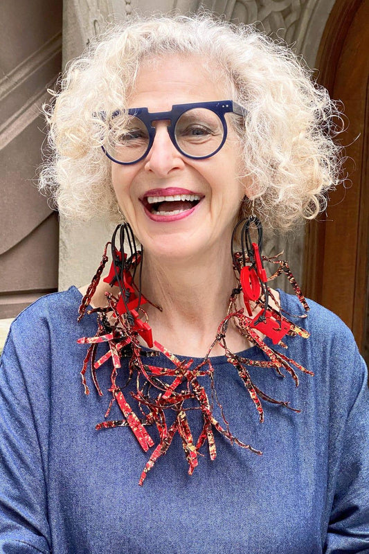 Happy older woman smiling into the camera. She is wearing a red, black and gold fleck necklace with love earrings. She is wearing a cotton denim dress.