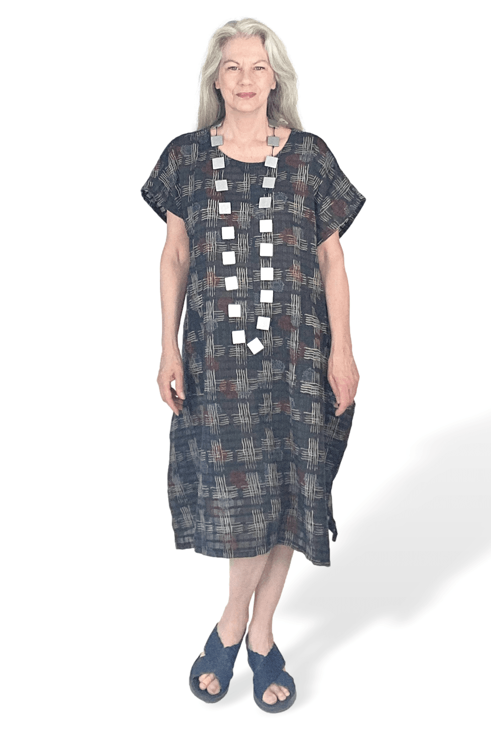 Woman wearing a short sleeve black loose fit dress with cross hatch patterns. She is wearing a long silver wooden square beaded necklace.