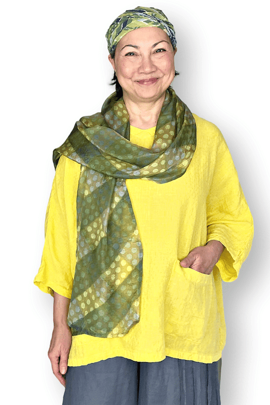 Smiling woman wearing a yellowish green polka dot silk scarf over a yellow linien full cut boxy top smiling.