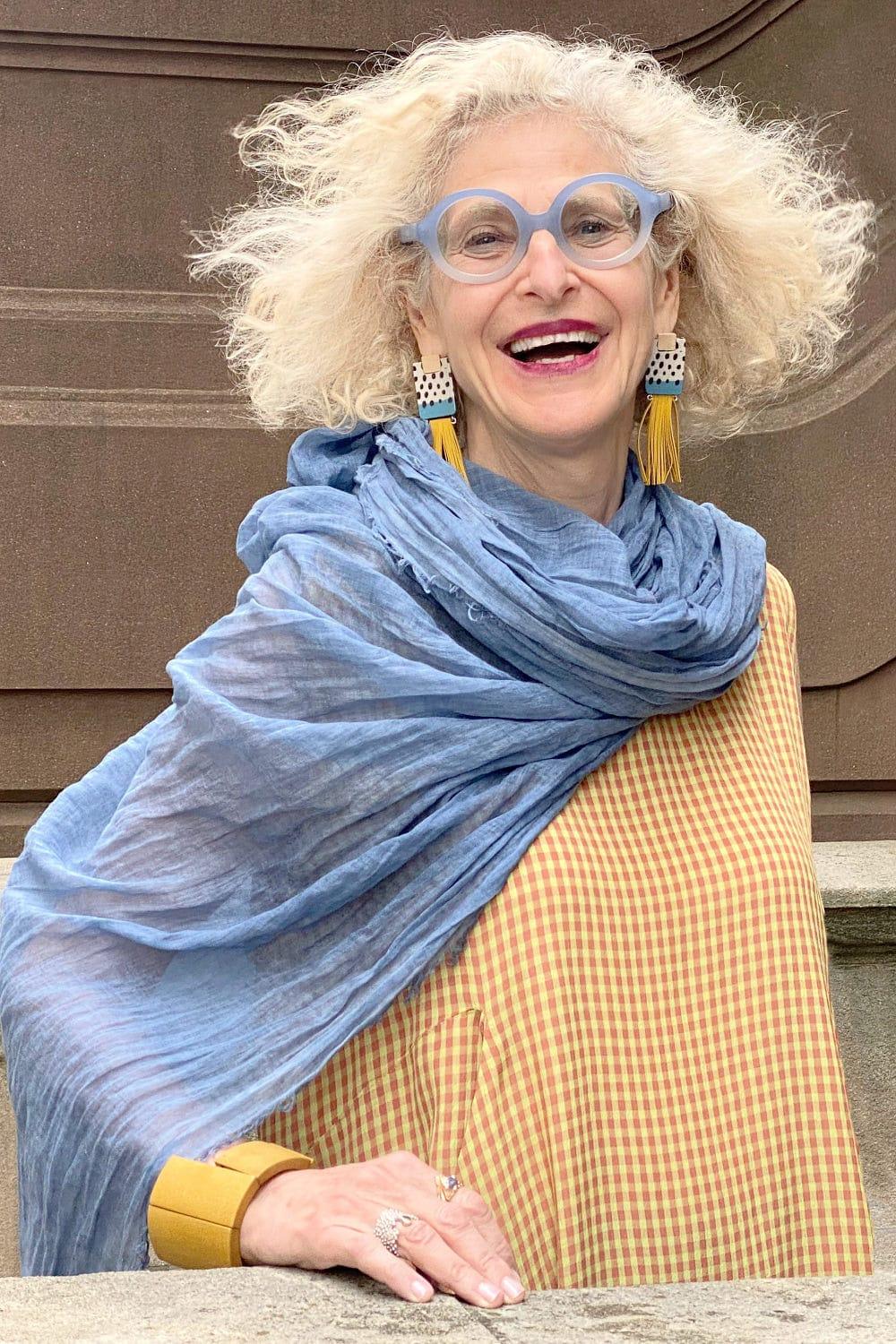 Woman wearing tassel earrings, blue scarf and a small check dress. She is wearing fun blue rimmed glasses and looks very stylish and happy.