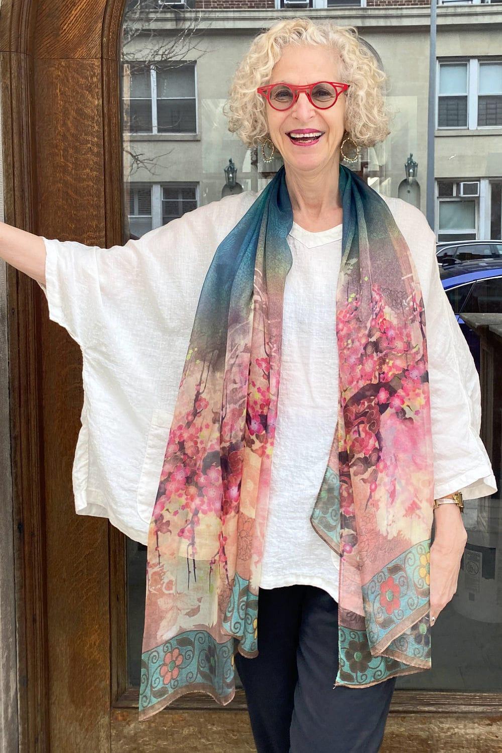 Sheer silk floral scarf colored with reds, pinks and teals, draped over the neck of a smiling woman over 60 years old. She is also wearing an oversized white linen top.