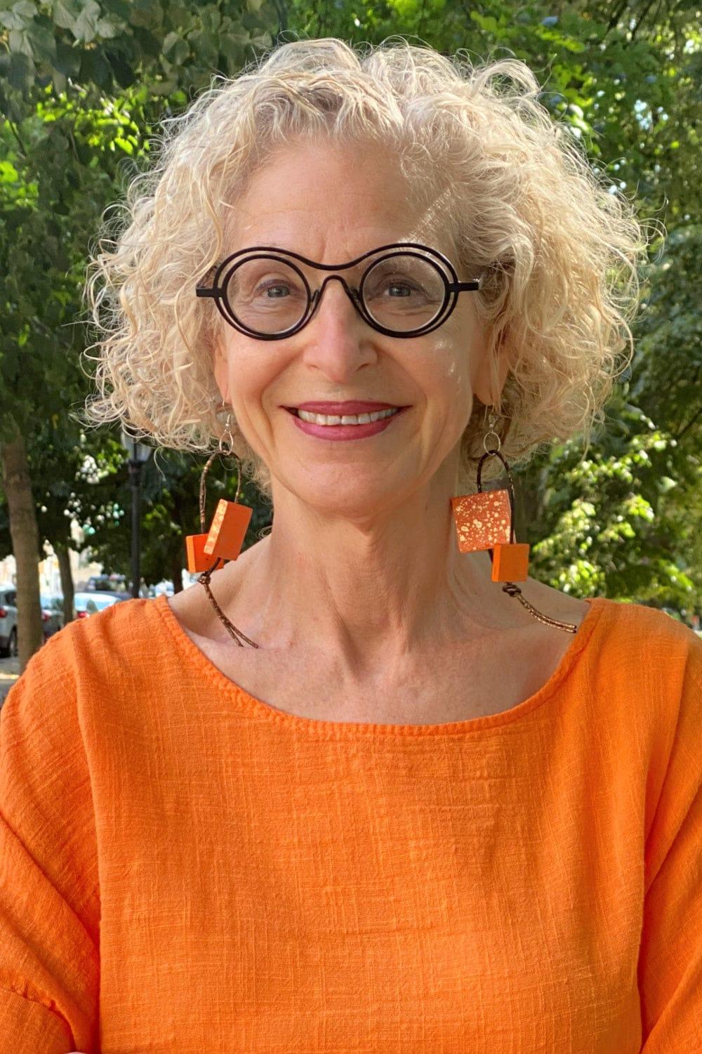 Smiling older woman wearing fun black rim glasses and orange dangle wooden earrings with an orang linen top.
