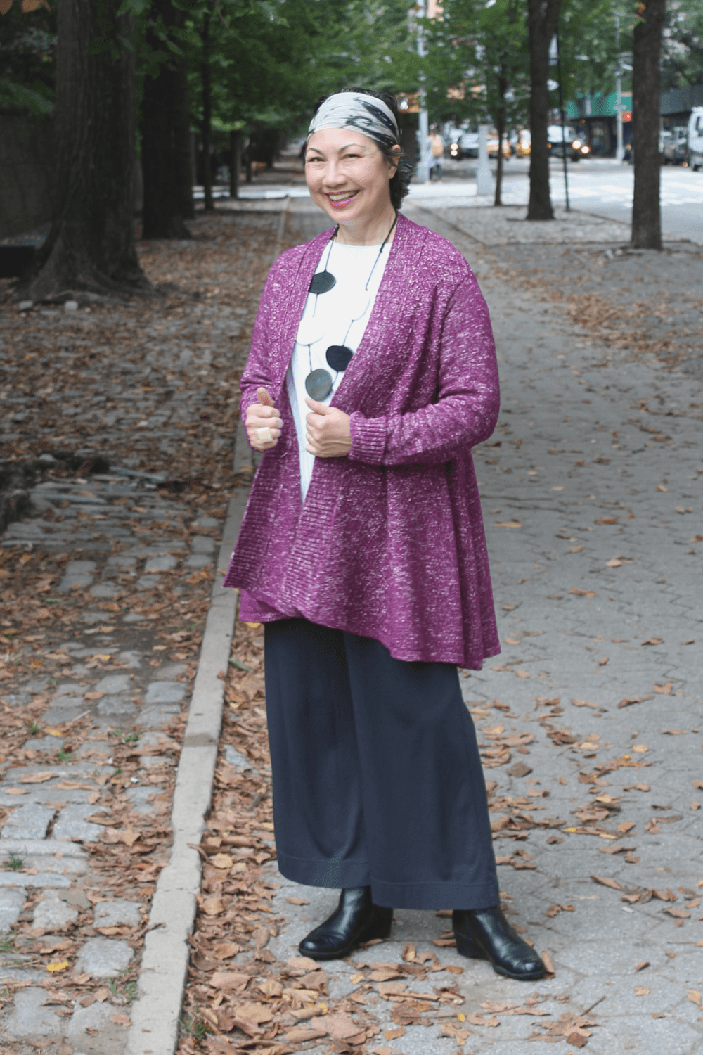 Woman standing in a park wearing a plum colored Cardigan with black full cut pants, a white top and a black and white necklace.