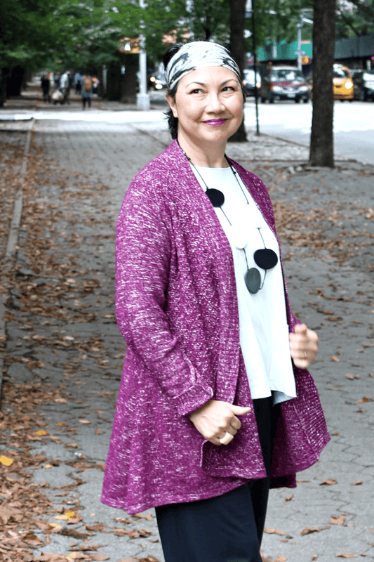 Woman standing in a park wearing a plum colored Cardigan with black full cut pants, a white top and a black and white necklace.