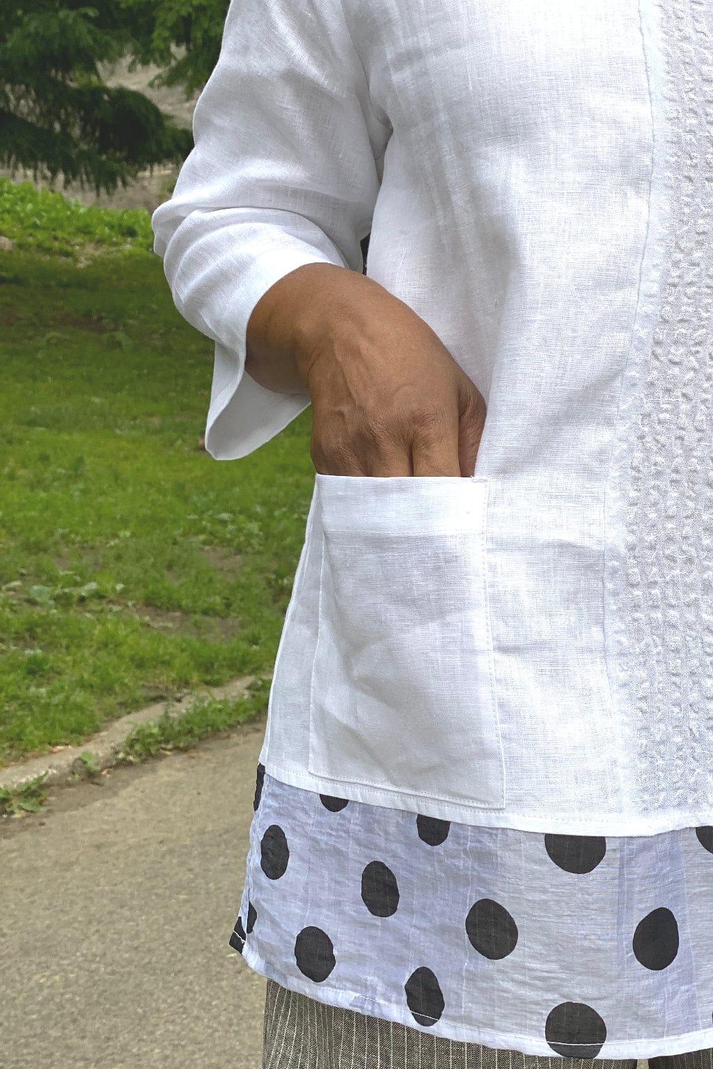 Close up of a white linen blouse with a front lower pocket. The top has a polka dot trim along the hemline.