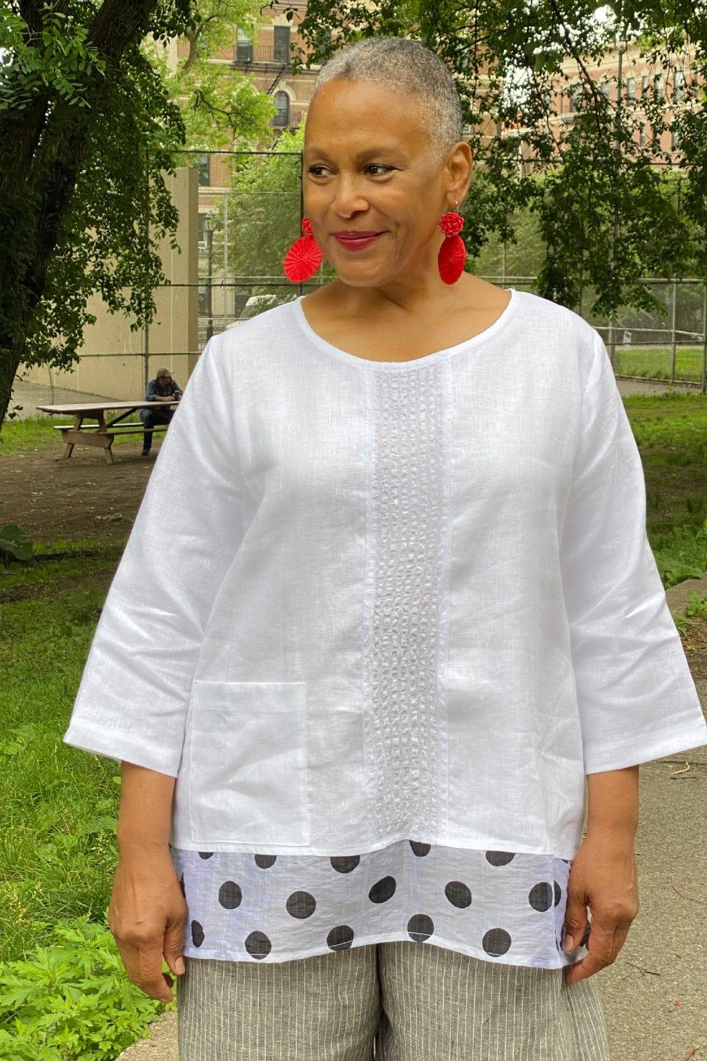 Woman with short cropped grey hair wearing a white linen top with a polka dot hem. She is wearing red woven earrings.