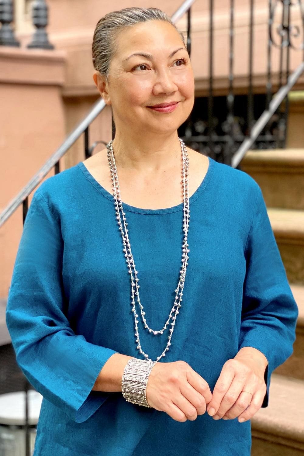 Women standing next to an outside staircase smiling and looking off into the distance. She is wearing a round neckline long sleeve blue linen top, a croquet beaded double strand necklace and a silver cuff bracelet. 